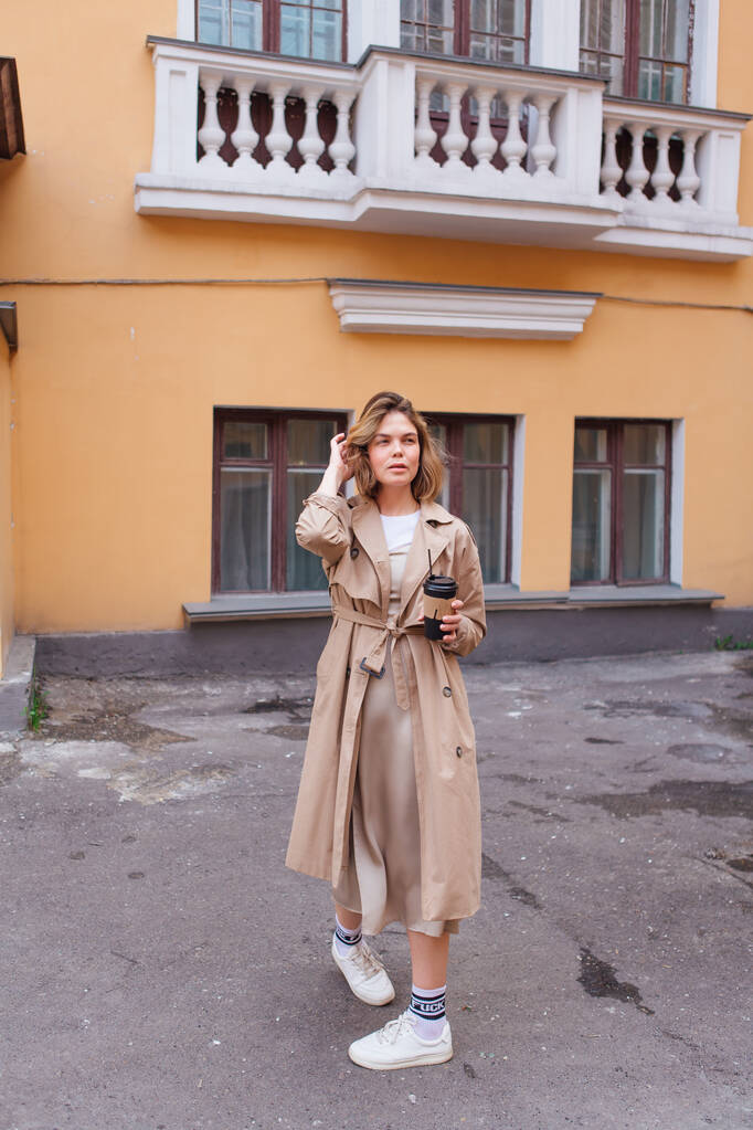 Genç milenyum kadını, sonbahar paltosu giymiş vahşi saçlarıyla elinde bir fincan kahveyle eski binanın yakınına gidiyor.. - Fotoğraf, Görsel