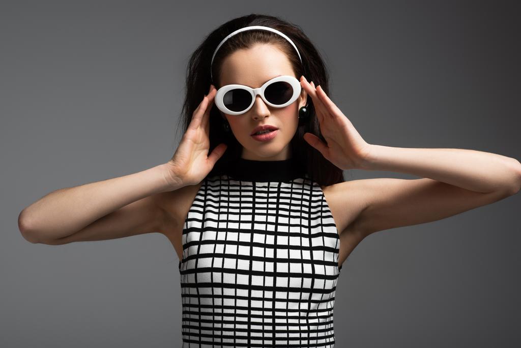 giovane donna in fascia regolando eleganti occhiali da sole isolati su grigio - Foto, immagini
