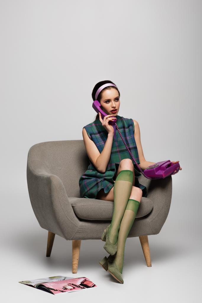 νεαρή γυναίκα μιλάει σε vintage τηλέφωνο, ενώ κάθεται στην πολυθρόνα κοντά περιοδικό στο πάτωμα σε γκρι - Φωτογραφία, εικόνα