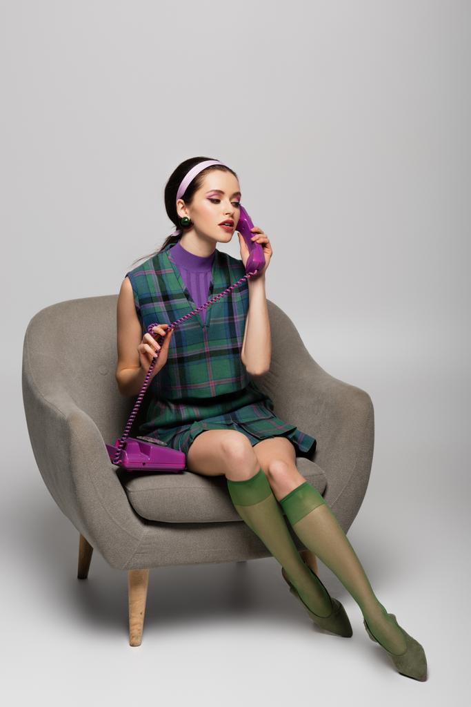 μελαχρινή νεαρή γυναίκα μιλάει vintage τηλέφωνο, ενώ κάθεται στην πολυθρόνα σε γκρι - Φωτογραφία, εικόνα