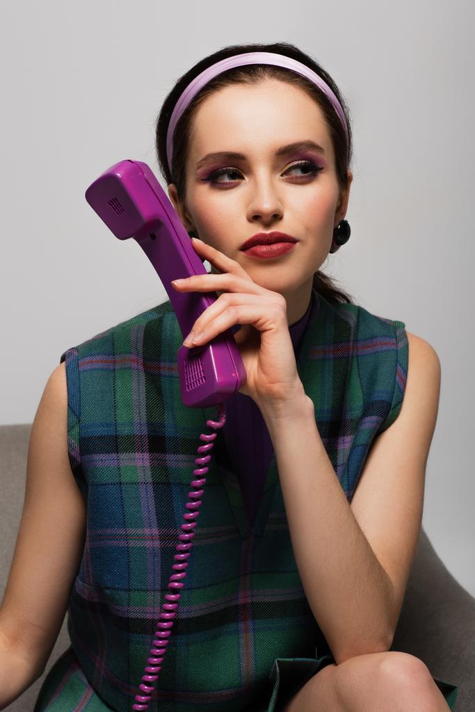 barna fiatal nő kezében retro telefon, miközben néz félre, és ül karosszékben elszigetelt szürke - Fotó, kép