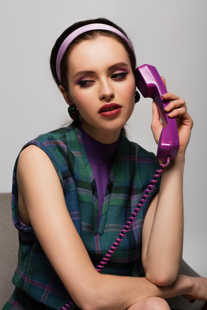 σκεπτική νεαρή γυναίκα μιλάει στο ρετρό τηλέφωνο, ενώ κάθεται στην πολυθρόνα απομονωμένη σε γκρι - Φωτογραφία, εικόνα