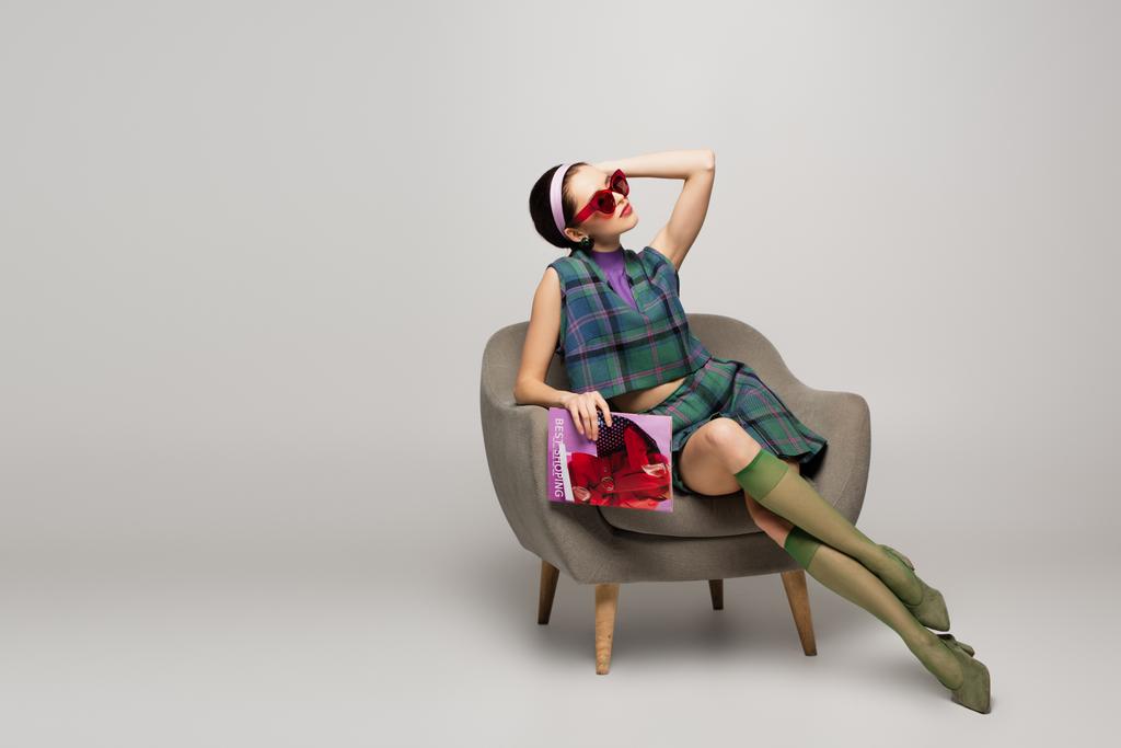 młoda kobieta w okularach przeciwsłonecznych gospodarstwa magazyn podczas pozowania i siedzi fotel na szarym - Zdjęcie, obraz
