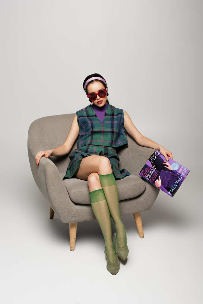 kobieta w okularach przeciwsłonecznych trzyma magazyn siedząc w fotelu na szarym - Zdjęcie, obraz
