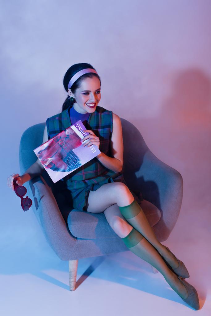 usmívající se mladá žena drží sluneční brýle a časopis, zatímco sedí v křesle na fialové - Fotografie, Obrázek
