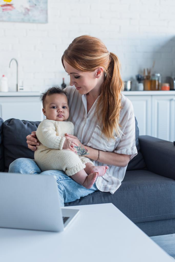 woman sitting on couch with infant african american daughter near laptop - Photo, Image