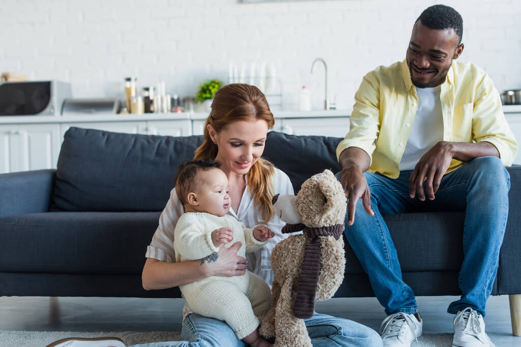glückliche Frau sitzt auf dem Boden mit kleiner Tochter und Teddybär neben lächelndem afrikanisch-amerikanischem Ehemann - Foto, Bild