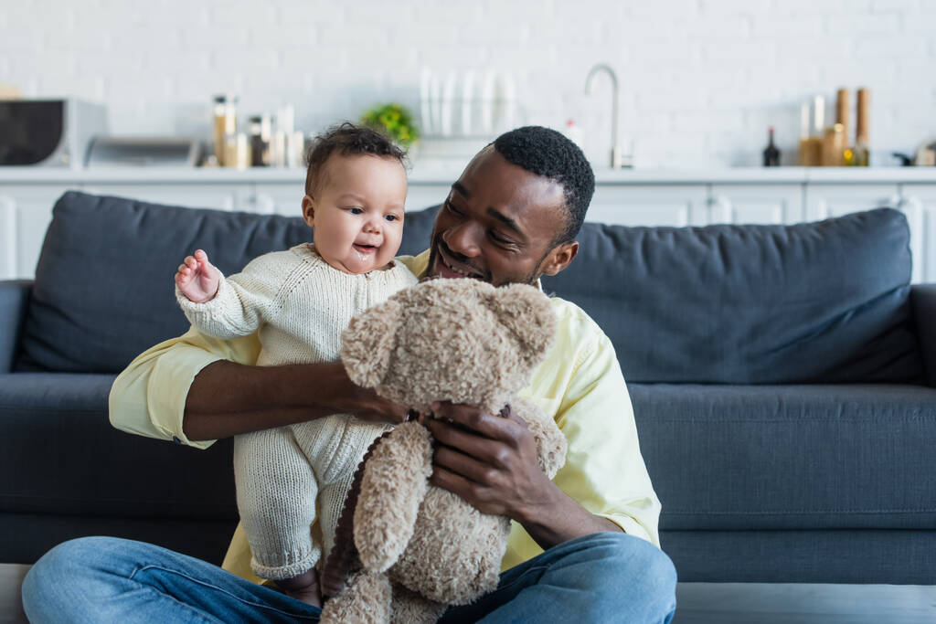 fröhliche afrikanisch-amerikanische Mann hält Teddybär beim Spielen mit Kleinkind Tochter - Foto, Bild