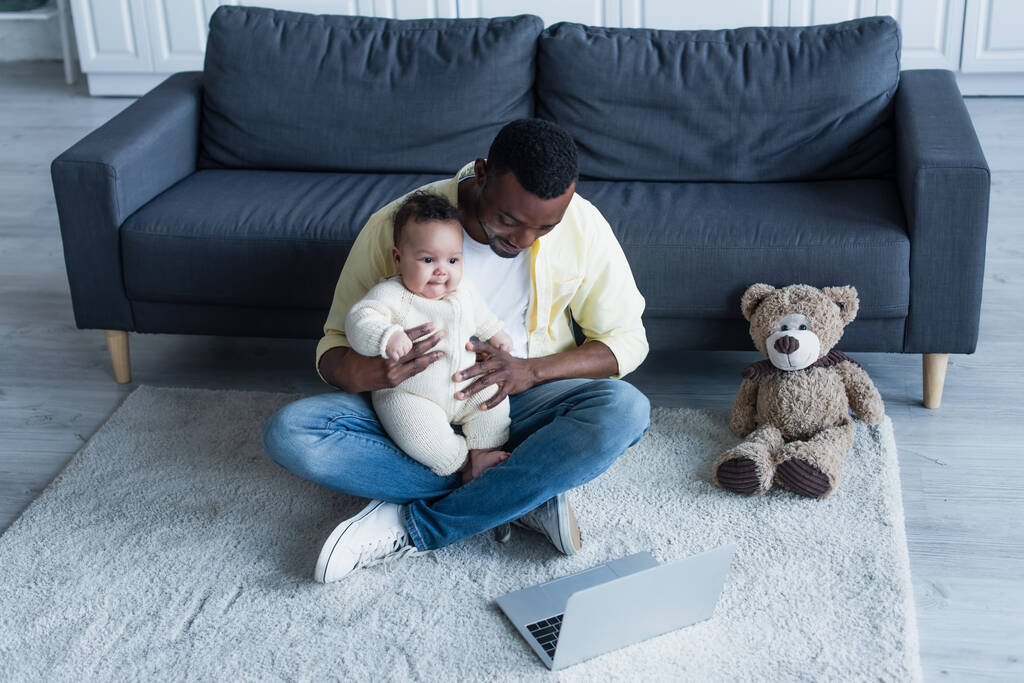 afro-amerikai férfi kezében baba, miközben ül a padlón mellett laptop és Teddy maci - Fotó, kép