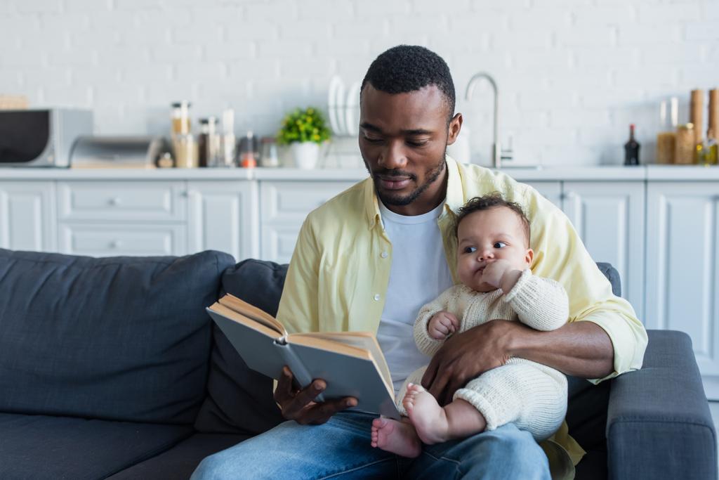afro-americano homem leitura livro para bebê menina no sofá na cozinha - Foto, Imagem
