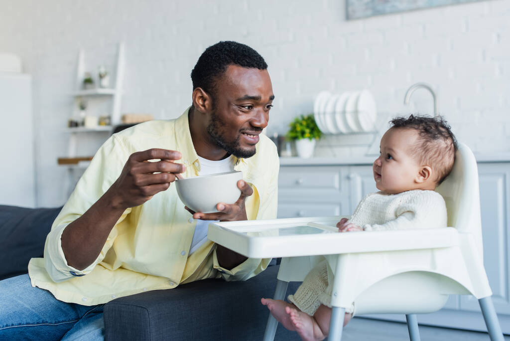 happy african american man holding bowl near infant kid sitting in baby chair - Photo, Image