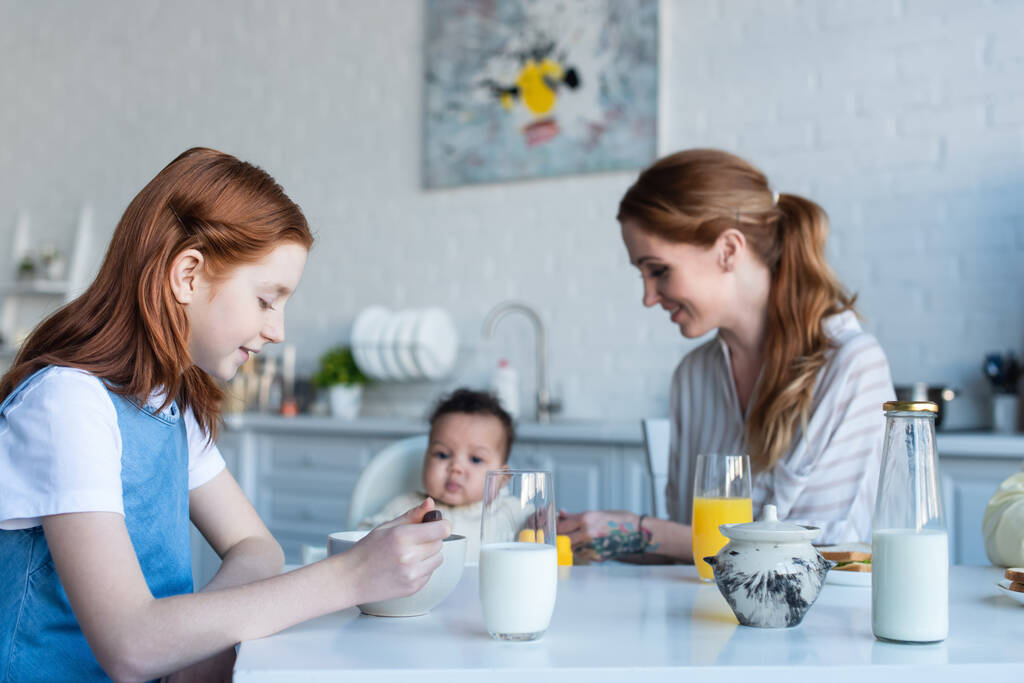 niña preadolescente desayunando cerca de la madre y el bebé afroamericano hermana - Foto, Imagen