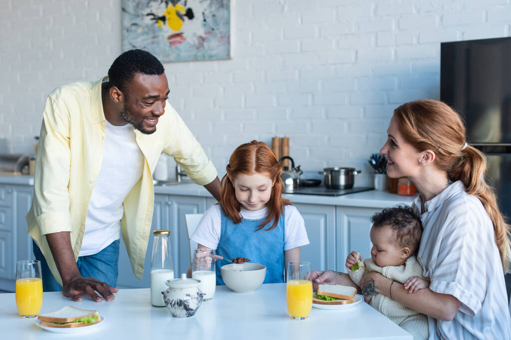 boldog afro-amerikai férfi áll közelében multietnikus lánya és felesége reggelizik a konyhában - Fotó, kép