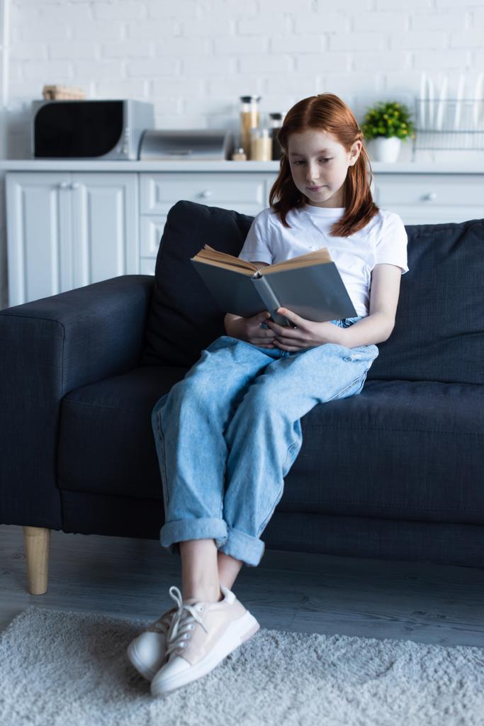 Evdeki kanepede kitap okuyan kızıl saçlı kızın tam boy görüntüsü. - Fotoğraf, Görsel