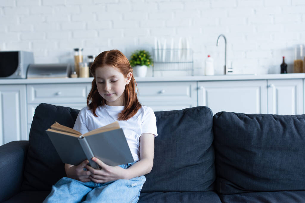 дошкольница читает книгу на диване на кухне - Фото, изображение