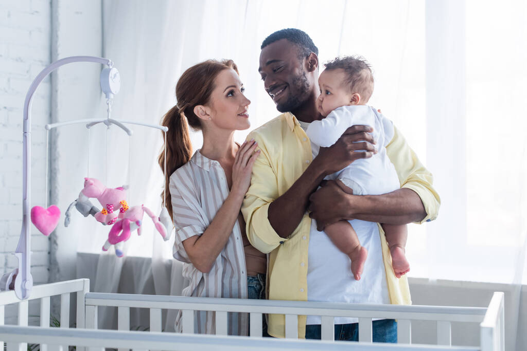 happy interracial couple with infant daughter standing near crib - Photo, Image