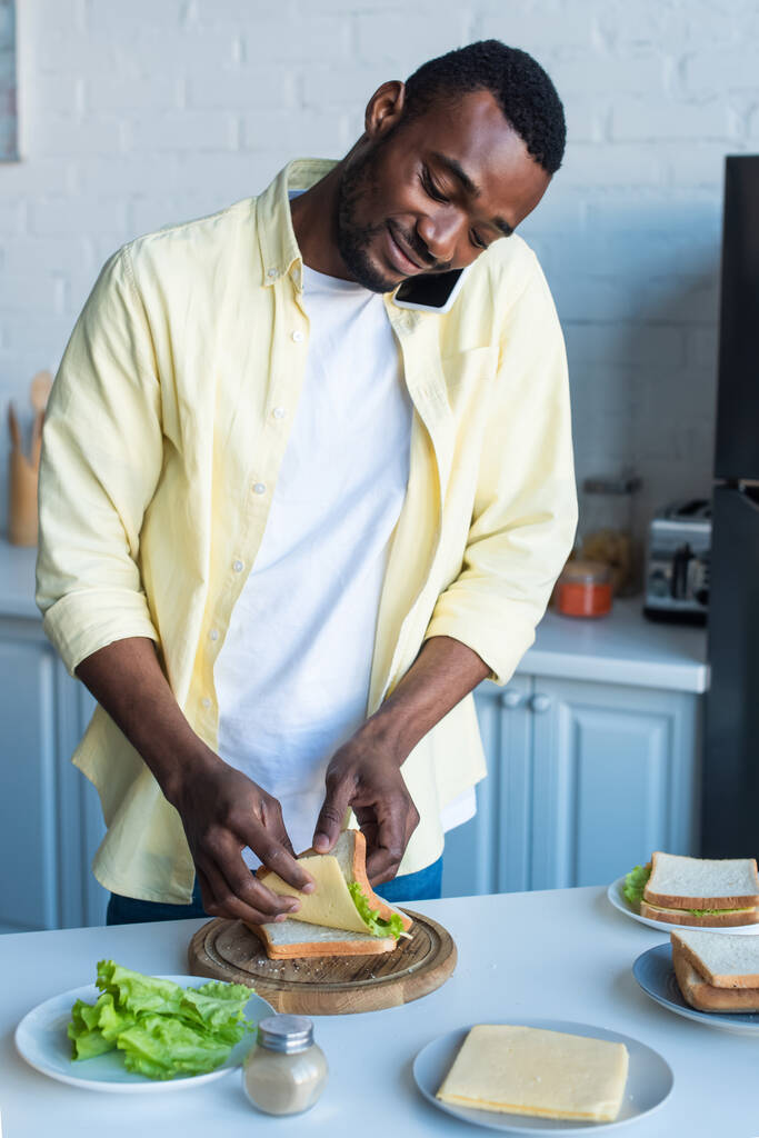 lachende Afrikaans-Amerikaanse man sandwiches maken en praten op smartphone - Foto, afbeelding