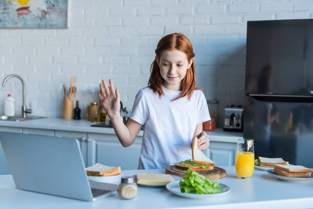 Дівчина махає рукою під час відеодзвінків і готує сніданок
 - Фото, зображення