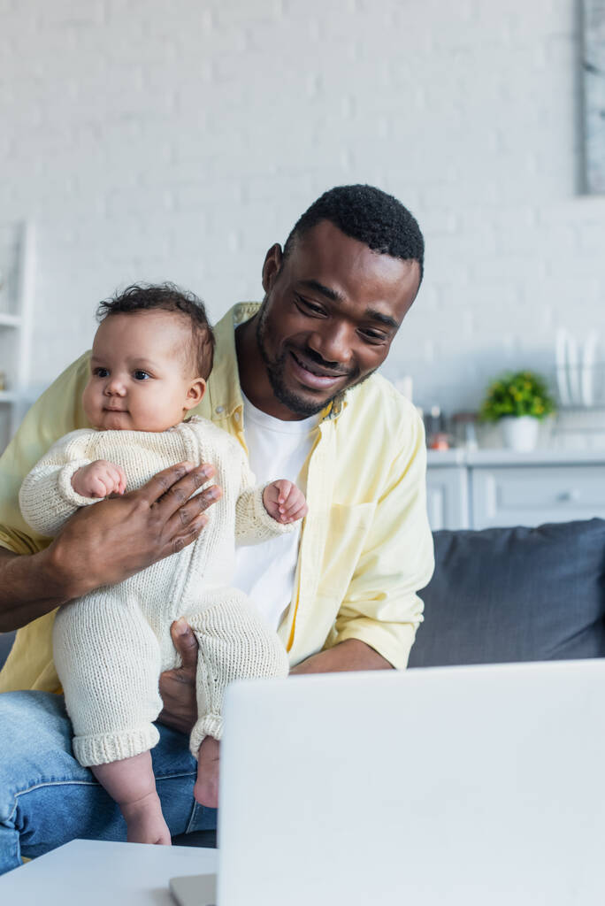 alegre afroamericano hombre sentado cerca borrosa portátil con bebé niño - Foto, Imagen