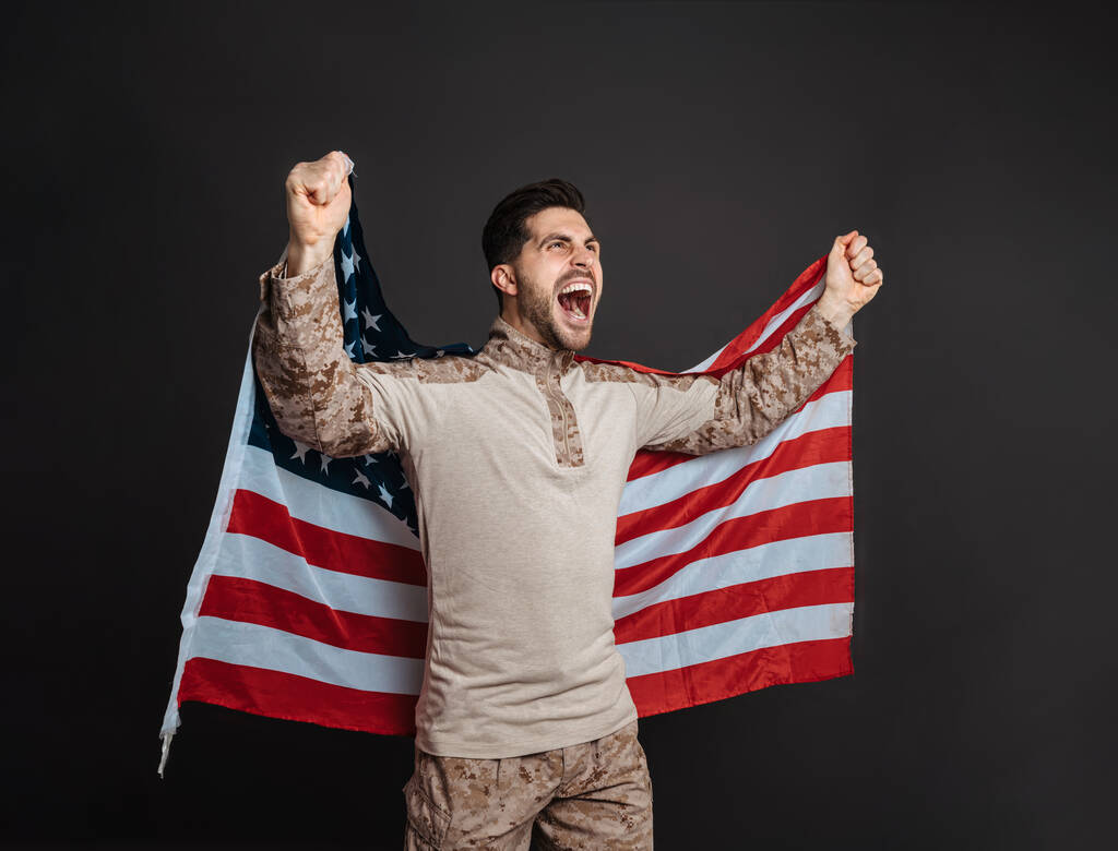 Aufgeregter Militärmann schreit, während er mit amerikanischer Flagge vor schwarzem Hintergrund posiert - Foto, Bild