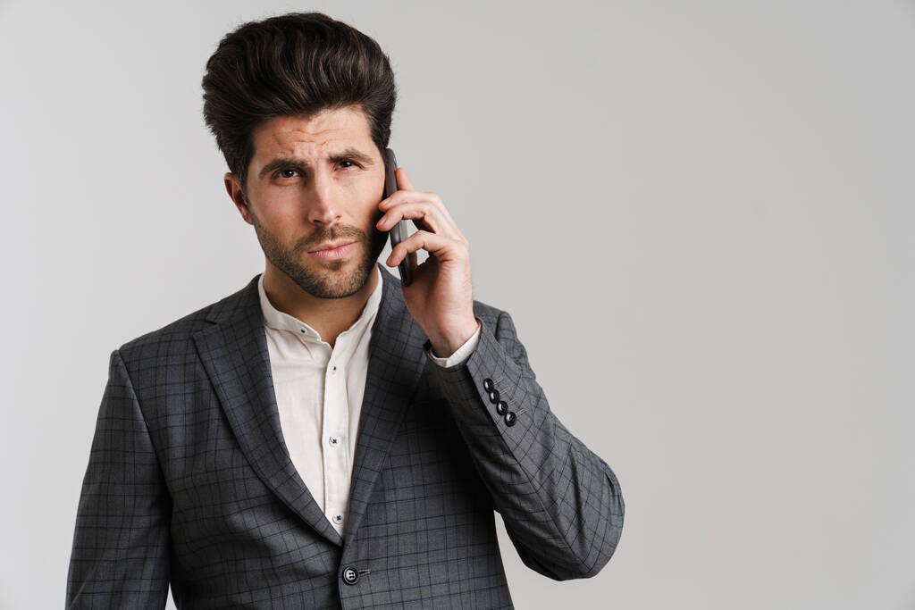 Vakava nuori mies takki poseeraa ja puhuu matkapuhelimella eristetty harmaalla taustalla - Valokuva, kuva