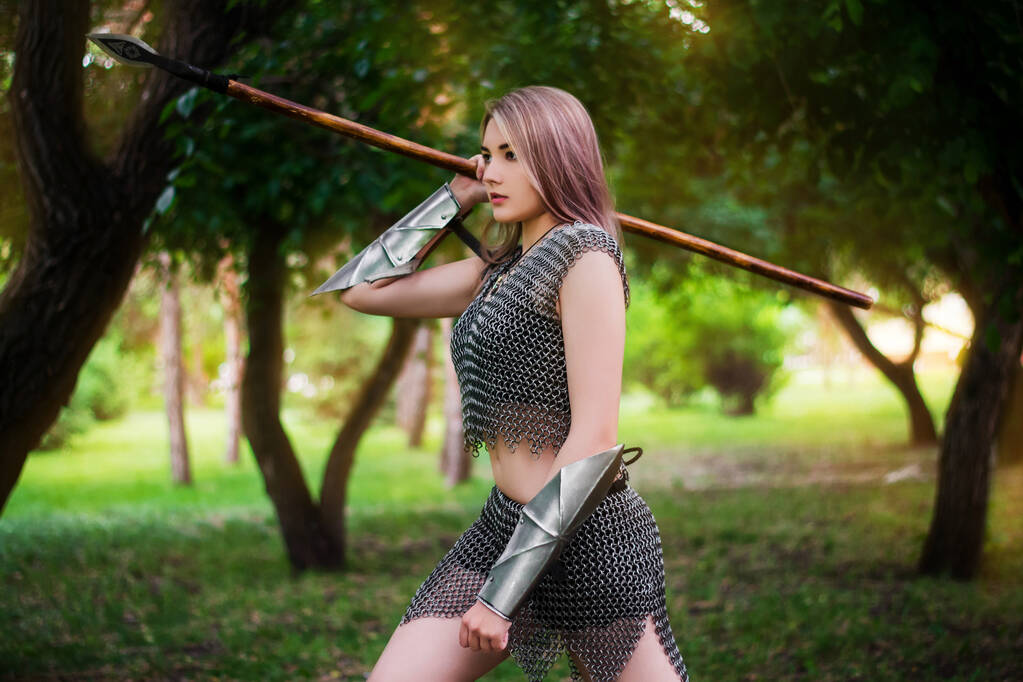Portrait of a young medieval woman warrior dressed in chain mail armor with a spear in her hands on the background of the forest. - Foto, Imagen