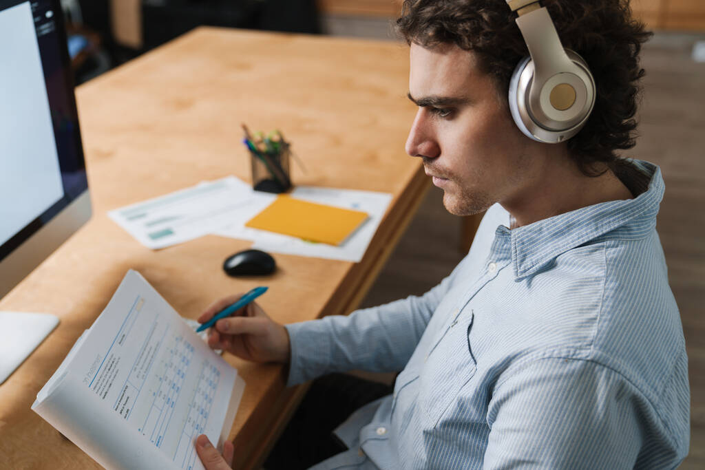 Καυκάσιος νεαρός επιχειρηματίας με ακουστικά που κρατάει έγγραφα ενώ εργάζεται στο γραφείο - Φωτογραφία, εικόνα