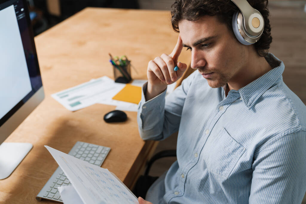 Hombre de negocios joven caucásico en auriculares con documentos en papel mientras trabaja en la oficina - Foto, Imagen