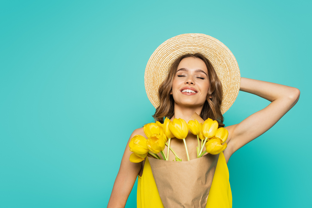 Веселая женщина в солнечной шляпе держит тюльпаны изолированы на синий  - Фото, изображение