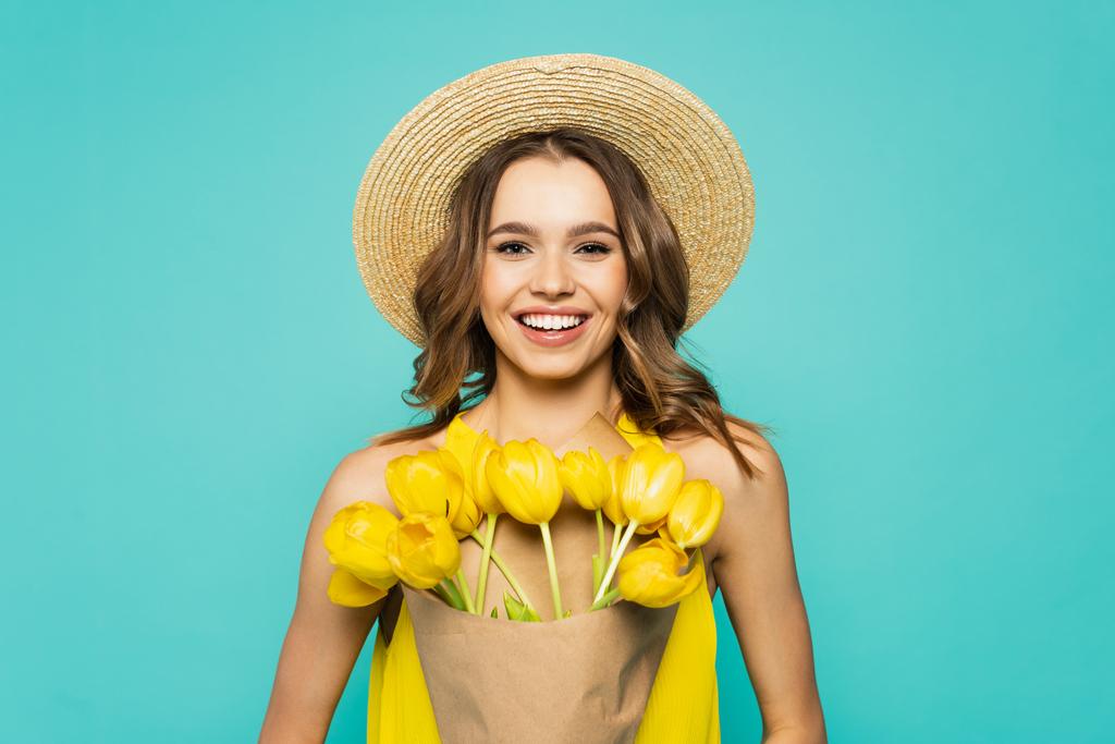 Hübsche Frau mit Sonnenhut hält Tulpen und lächelt isoliert auf blauem Grund  - Foto, Bild