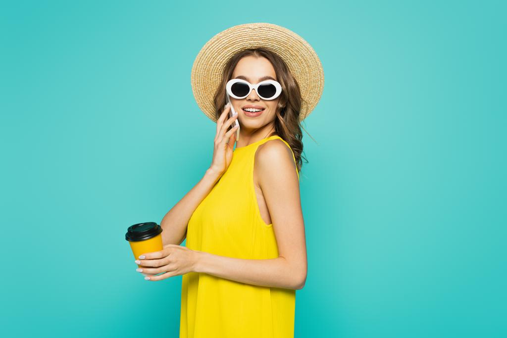 Femme souriante en chapeau de soleil parlant sur smartphone et tenant du café pour aller isolé sur bleu  - Photo, image