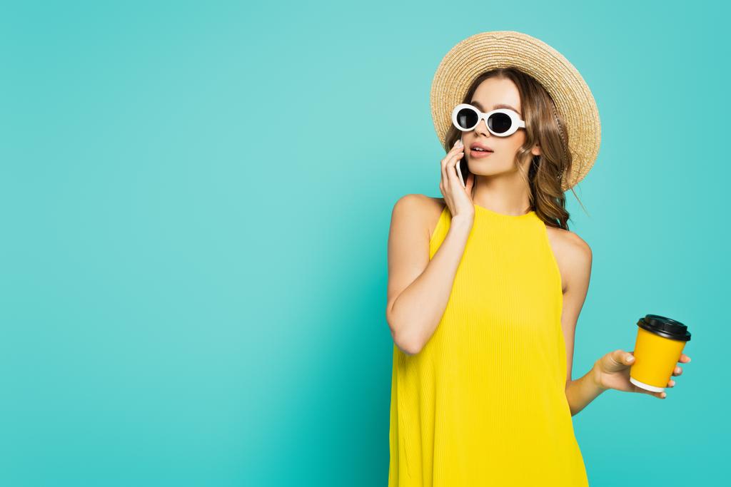 Nuori nainen aurinkolasit ja aurinko hattu puhuu matkapuhelimeen ja pitämällä paperimuki eristetty sininen  - Valokuva, kuva