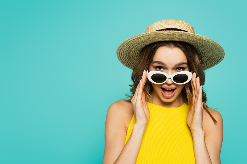 Femme excitée en chapeau de paille tenant des lunettes de soleil isolées sur bleu  - Photo, image