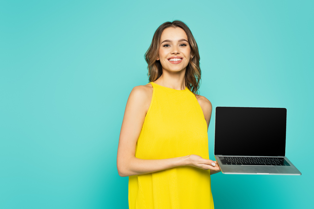 Fröhliche Frau hält Laptop in der Hand und blickt in die Kamera auf blauem Hintergrund  - Foto, Bild