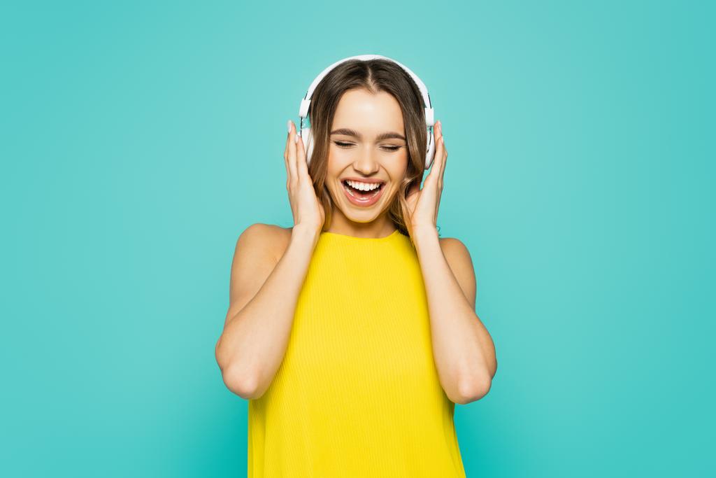 Positive Frau in gelbem Kleid hört Musik in Kopfhörern isoliert auf blauem Grund  - Foto, Bild