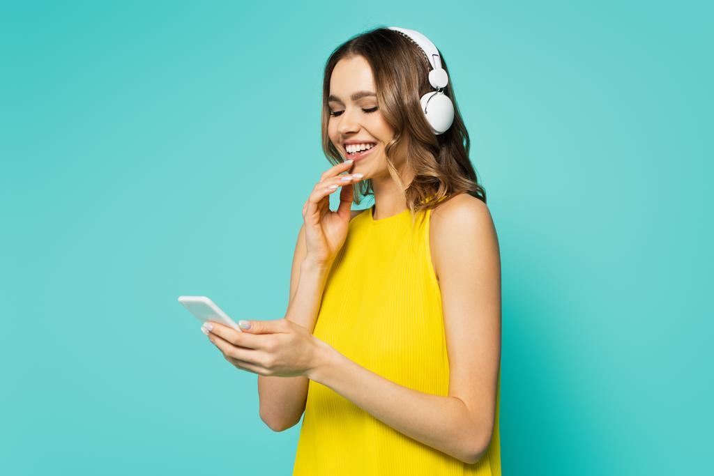 Mulher sorridente em fones de ouvido usando smartphone isolado em azul  - Foto, Imagem
