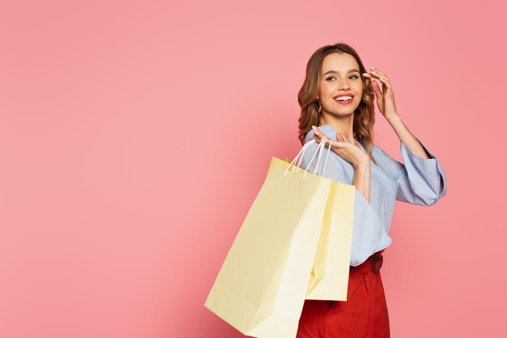 Mujer joven sosteniendo bolsas de compras y mirando hacia otro lado aislado en rosa  - Foto, Imagen