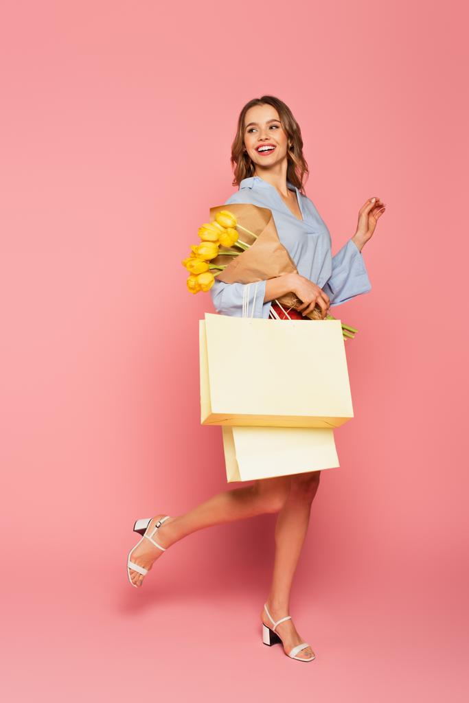 Mujer alegre sosteniendo ramo y bolsas de compras sobre fondo rosa  - Foto, imagen