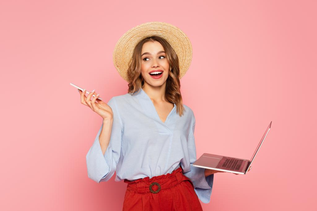 Smiling woman in sun hat holding smartphone and laptop isolated on pink  - Photo, Image