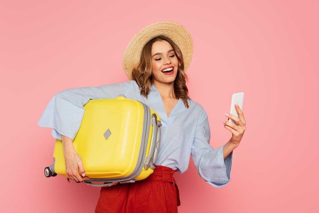 Femme souriante avec valise utilisant un smartphone isolé sur rose  - Photo, image