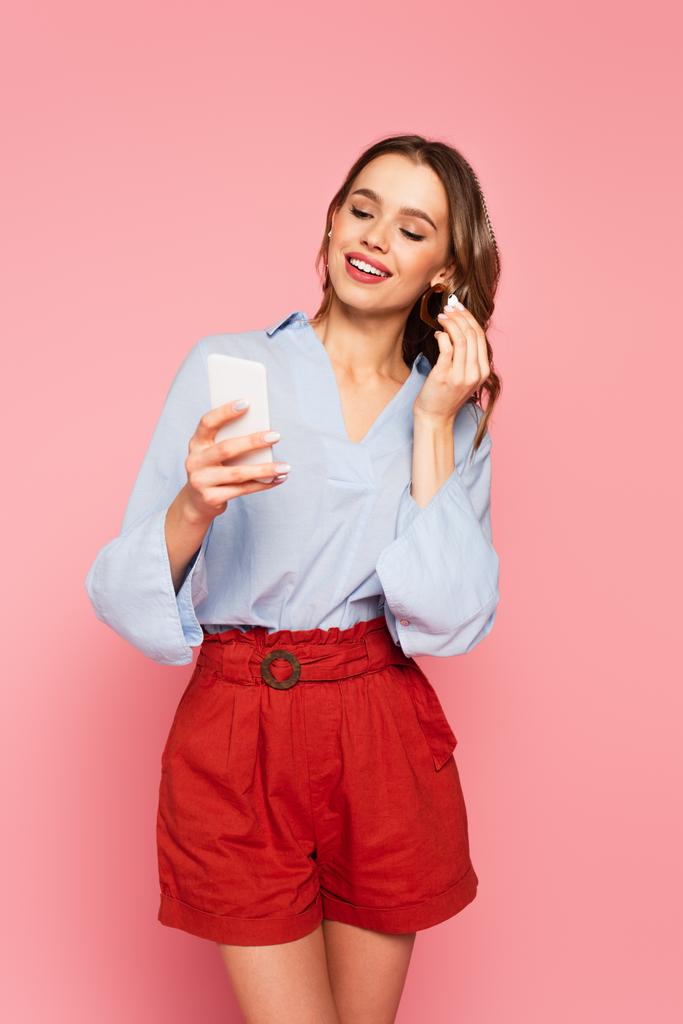 Красива жінка посміхається, тримаючи навушники та смартфон ізольовано на рожевому
  - Фото, зображення