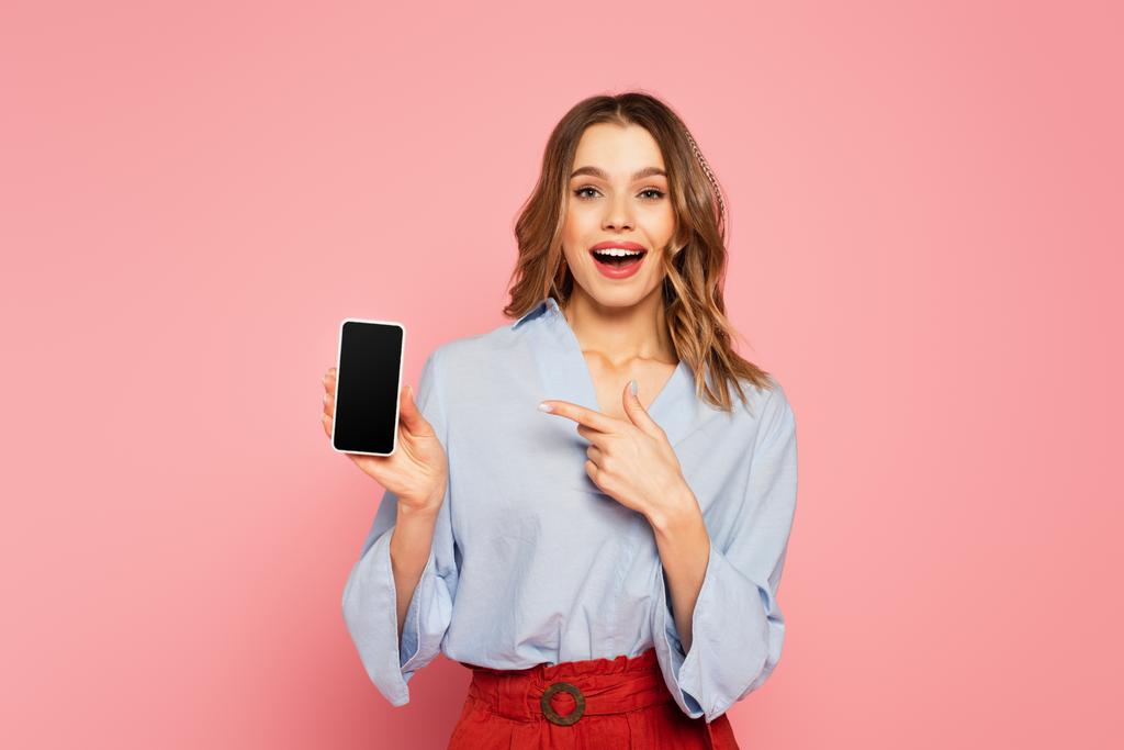 Aufgeregte Frau zeigt auf Smartphone mit leerem Bildschirm auf rosa Hintergrund  - Foto, Bild