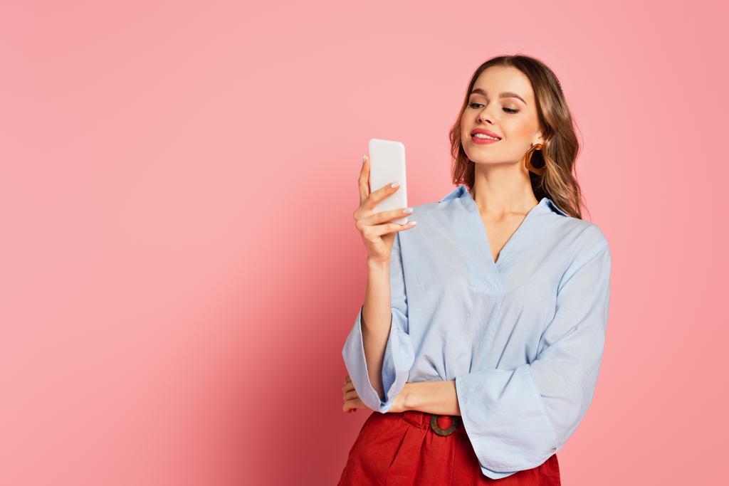 Stílusos nő használ okostelefon és mosolygós rózsaszín háttér  - Fotó, kép