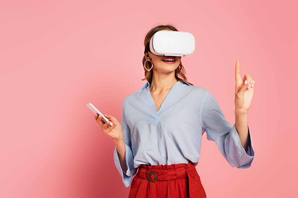 Smiling woman in vr headset pointing with finger on pink background  - Photo, Image
