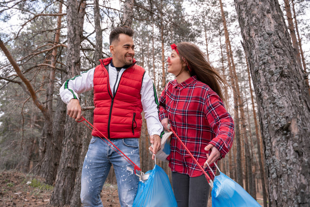 happy couple with trash bags picking up rubbish with grabber tools in forest  - Photo, Image