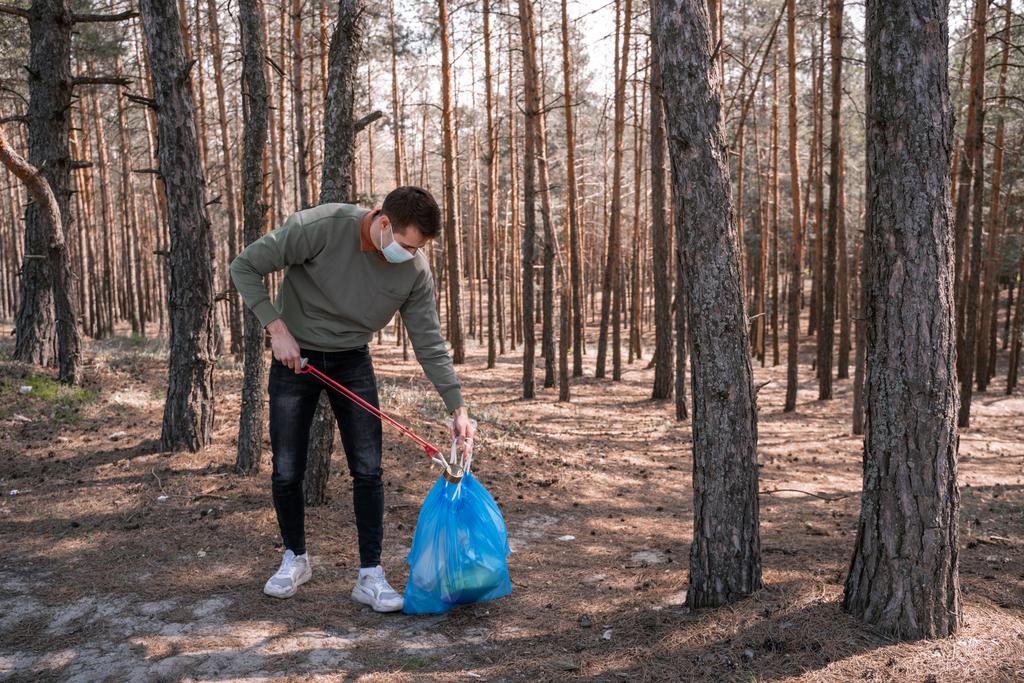 Medikal maskeli bir adam ormanda çöpün yanında bir alet tutuyor.  - Fotoğraf, Görsel