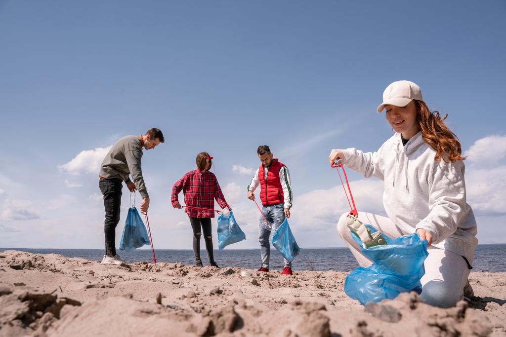 femme heureuse tenant sac poubelle et ramassant des ordures sur le sable près du groupe de bénévoles  - Photo, image