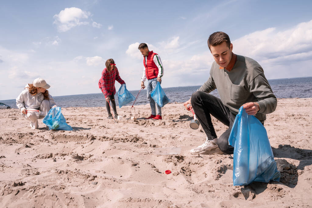 ゴミ袋を持ってボランティアのグループの近くの砂の上にゴミを集める若者は  - 写真・画像