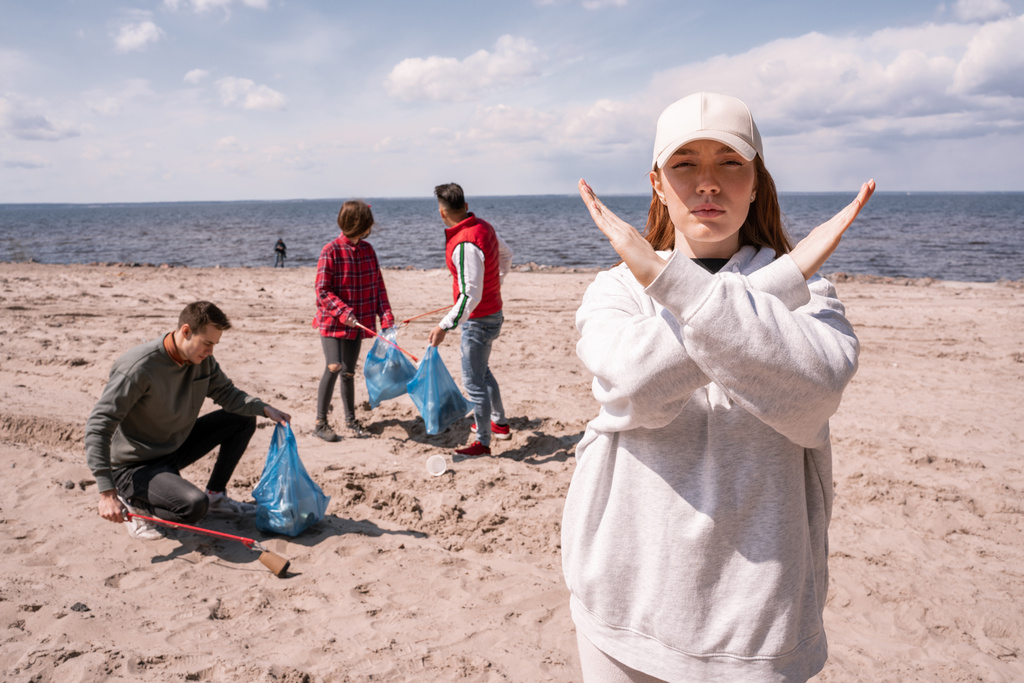 ゴミ拾いのボランティアグループの近くでストップジェスチャーを見せるキャップの若い女性生態学の概念  - 写真・画像