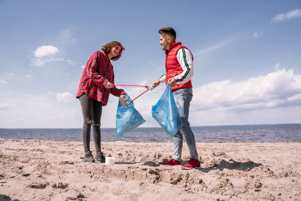 feliz pareja con bolsas de basura y agarradores recogiendo basura en la arena  - Foto, imagen